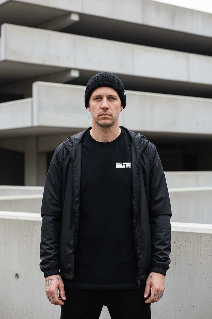 30 something man stands in front of cold brutalist architecture wearing the "borders are hostile" t-shirt